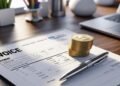 A printed invoice lies on a desk alongside a pen. Nearby is a stack of gold coins with a dollar sign. The background shows a laptop and stationery, indicating a work or financial setting.